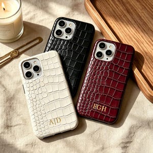 以下が含まれることがあります： クロコダイル柄の型押しが施されたスマートフォンケース3点。白のケースには金色のモノグラム「A|D」、黒のケース、そして濃い赤のケースには金色のモノグラム「FGH」が刻印されています。各ケースの背面には3眼カメラ用の切り抜きがあります。