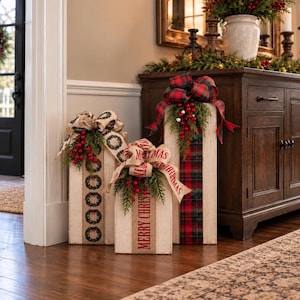 May include: Three rectangular wooden blocks painted white with distressed edges, decorated as gift boxes. Each features a burlap or plaid ribbon bow, green pine sprigs, red berries, and text including Merry Christmas.