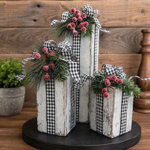 May include: Three rectangular wooden blocks painted with a distressed white finish, each wrapped in black and white gingham ribbon. The tops are decorated with artificial pine needles, red berries, and matching gingham bows.