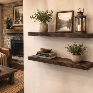 May include: Two dark brown wooden floating shelves mounted on a white wall. The top shelf holds a ceramic vase with greenery, a framed landscape print, and a metal lantern. The bottom shelf holds a stack of books, a wooden bowl, and a potted plant.