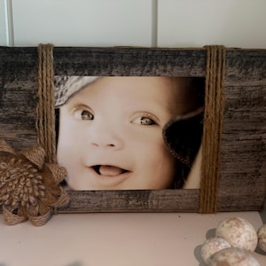 May include: A rectangular picture frame crafted from weathered gray wood, featuring vertical wraps of natural brown jute twine on the left and right sides. A decorative flower made of jute fabric is attached to the bottom left corner.