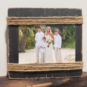 May include: A rectangular black wooden picture frame with distressed edges, featuring thick natural jute twine wrapped horizontally around the top and bottom sections. The frame holds a photograph of a person in a white dress and three others.