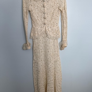 May include: A long-sleeved, floor-length cream lace dress featuring a fitted bodice with a row of four fabric-covered buttons down the front, a pointed collar with ruffled edges, and matching ruffled cuffs at the end of each long sleeve.