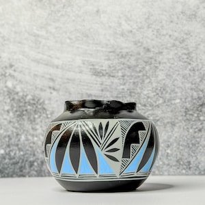 May include: A small, rounded ceramic pot featuring a black base colour with intricate geometric patterns in light blue, white, and black. The design includes repeating fan-like shapes and linear details around the body of the vessel.