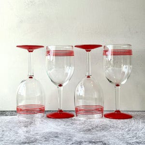 May include: Four clear glass wine glasses with red accents. Two glasses are upright, and two are inverted. Each glass features a series of thin, horizontal red stripes around the bowl. The inverted glasses have red bases, while the upright glasses have red stems and bases.