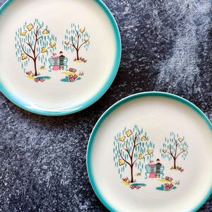 Peut inclure: Deux assiettes rondes en céramique avec un centre crème et un bord turquoise vif. Le centre de chaque assiette présente une illustration graphique de deux personnages assis sur un banc turquoise entre deux arbres aux fleurs jaunes et feuilles vertes.