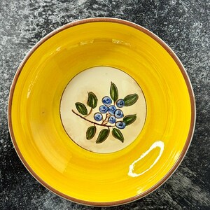 May include: A round ceramic bowl featuring a bright yellow interior rim and a cream-colored center base. The center of the bowl is decorated with a hand-painted illustration of a cluster of blue blueberries on a brown stem with green leaves.