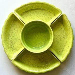 Puede incluir: Un juego de servir de cerámica en color verde lima que consta de un cuenco circular central rodeado por cuatro platos separados en forma de abanico. Cada pieza presenta un patrón moteado con manchas verde oscuro y un borde festoneado.