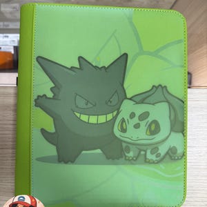 May include: A lime green rectangular binder featuring a graphic print of the dark purple Gengar and the light green Bulbasaur characters. The cover has a textured finish with stitched edges and a subtle leaf pattern background design.