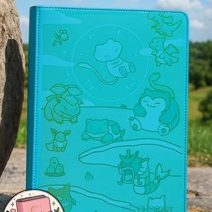 Peut inclure: Un classeur turquoise vif orné d'un motif gravé représentant diverses créatures de dessin animé, dont une figure flottante centrale, un grand personnage assis et une créature aquatique. Le texte PHOENIX SHIELD apparaît en bas à droite.