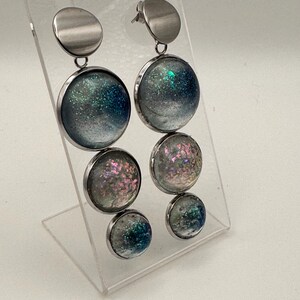 Può includere: Un paio di orecchini pendenti con tre ciondoli circolari in resina di diverse dimensioni sospesi da un perno in metallo argentato. Gli elementi in resina presentano una sfumatura di particelle glitter blu, turchesi e rosa iridescente.