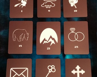 Puede incluir: Nueve cartas con ilustraciones blancas sobre un fondo marrón. Las cartas están numeradas del 1 al 36. Las ilustraciones incluyen una bruja en una escoba, una nube con gotas de lluvia, una parca, un oso, una cordillera, dos anillos, un sobre, una llave y una cruz.