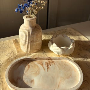 Op de afbeelding: Een beige gemarmerde vaas met verticale ribbels met blauwe en crèmekleurige droogbloemen, een witte lotusvormige kom en een ovaal beige gemarmerd dienblad. De items zijn samen gerangschikt en tonen gladde texturen en natuurlijke patronen.