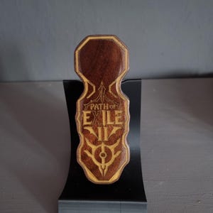Könnte beinhalten: Eine dunkelbraune Holzplakette mit glatter Oberfläche und hellem Rand. Die Plakette zeigt den eingravierten Text "PATH OF EXILE II" und ein stilisiertes kreisförmiges Emblem unter dem Schriftzug, auf einem schwarzen, geschwungenen Ständer.