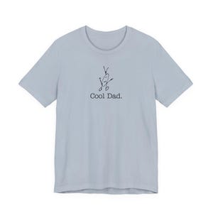 May include: A light blue t-shirt with a crew neck and short sleeves. The shirt features a whimsical black line drawing of a figure with the text "Cool Dad."