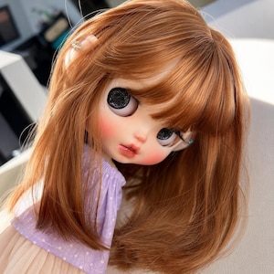 May include: A doll with shoulder-length auburn hair, bangs, and large, dark, glittery eyes. The doll has a pale complexion with rosy cheeks and pink lips. It is wearing a light purple, short-sleeved top with a white polka dot pattern.
