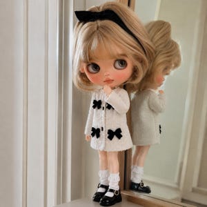 May include: A doll with blonde hair styled in a bob, wearing a black headband, a white textured coat with four black bows, white ruffled socks, and black Mary Jane shoes. The doll has large eyes and is positioned in front of a mirror reflection.