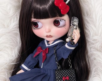 May include: A doll with long dark hair, red eyes, and a red bow hair clip. It wears a navy blue sailor-style dress with a red bow at the collar. The doll holds a clear, glittery flip phone accessory and carries a small black quilted handbag.