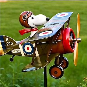 May include: A metal garden spinner featuring a white cartoon dog wearing a brown aviator helmet and red scarf, piloting a green biplane. The plane has a red nose, yellow propeller, and blue and white roundel markings with the text B 7270 on the tail.