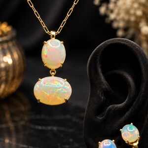 May include: A gold necklace featuring two stacked oval opal cabochons and a pair of matching gold opal stud earrings. Each opal displays a milky white base with iridescent flashes of red, green, and blue colour, secured in simple gold prong settings.