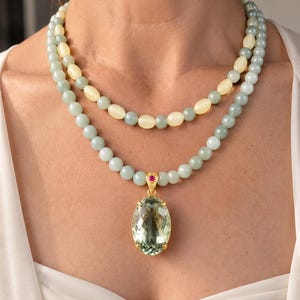 Op de afbeelding: Een dubbelstrengs ketting met lichtgroene en crèmekleurige kralen. De onderste streng bevat een grote, ovaal geslepen, lichtgroene edelsteen hanger gezet in goud, geaccentueerd met een kleine, felroze steen aan de gouden hangerhouder.