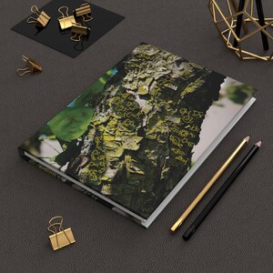 May include: A rectangular hardcover notebook featuring a photographic print of tree bark with green moss and yellow line-art drawings of stick figures and shapes. The notebook is positioned on a dark textured surface with gold and black pencils.