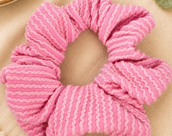 May include: A circular hair scrunchie featuring a ribbed texture with alternating stripes of bright pink and light pink. The fabric is gathered into a ruffled, voluminous shape, creating a soft, flexible accessory for securing hair in place.