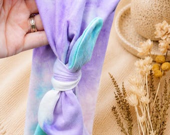 May include: A wide fabric headband featuring a tie-dye pattern in shades of purple, blue, and white. The headband has a central knotted detail that creates a bow-like appearance on the front, with soft, flexible material throughout the design.