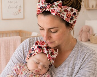May include: Two matching headbands featuring a white base with a vibrant red, pink, and dark floral pattern. Each headband is wide and includes a prominent knotted bow detail at the center. The fabric has a textured appearance.