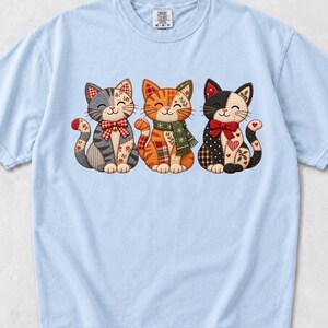 Könnte beinhalten: Ein hellblaues Kurzarm-T-Shirt mit einer Grafik von drei Katzen im Patchwork-Stil. Die linke Katze ist grau, die mittlere Katze ist orange und die rechte Katze ist schwarz. Sie tragen festliche Accessoires wie Schleifen und einen Schal.