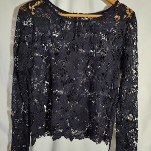 May include: A long-sleeved black lace top featuring an intricate floral pattern throughout the fabric. The garment has a round neckline and a scalloped hemline, with a visible zipper closure at the back of the neck. The lace is semi-sheer.