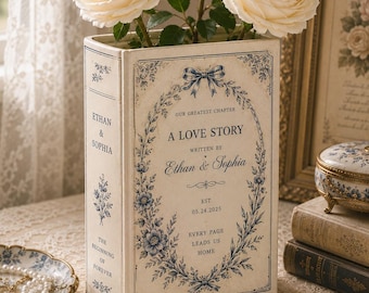 May include: A rectangular off-white book-shaped vase featuring blue floral illustrations and text that reads A LOVE STORY, WRITTEN BY Ethan & Sophia, EST. 05.24.2025, EVERY PAGE LEADS US HOME, and THE BEGINNING OF FOREVER, holding three cream-coloured roses.