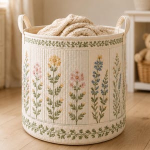 May include: A cylindrical fabric storage basket with two side handles, featuring a cream-coloured quilted exterior decorated with vertical panels of embroidered wildflowers in shades of yellow, pink, and blue, and a green vine border.