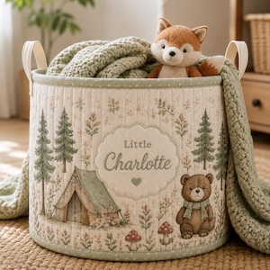 Puede incluir: Una cesta de almacenamiento cilíndrica de tela acolchada con un estampado de bosque que incluye pinos, una tienda de campaña, setas y un oso pardo. En el centro, una etiqueta festoneada muestra el texto Little Charlotte y un corazón.