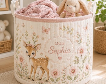 May include: A cylindrical fabric storage basket with a quilted cream exterior featuring a printed fawn, pink flowers, and butterflies. The basket displays the text Little Sophia in a scalloped frame, contains a pink knit blanket and a plush bunny.