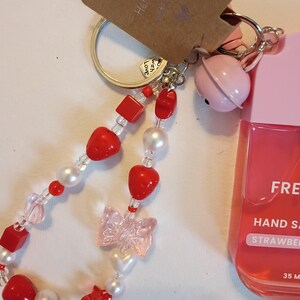 Pode incluir: Um chaveiro artesanal com uma tira de contas com contas vermelhas em forma de coração e quadradas, pérolas brancas e um laço rosa transparente. Uma campainha rosa e um frasco de desinfetante para as mãos com aroma de morango estão presos. A etiqueta diz "Handmade with Love."