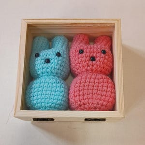 May include: Two small crocheted bunnies, one light blue and one pink, are placed inside a square, light-colored wooden box. Each bunny features a textured yarn surface, two upright ears, and three small black beads for eyes and a nose.