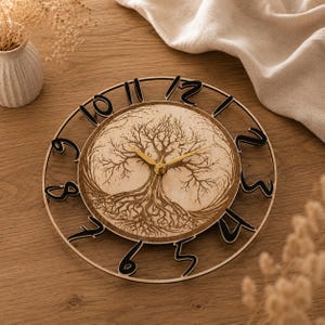 Può includere: Un orologio da parete circolare con un pannello centrale in legno chiaro e un disegno dell'albero della vita inciso al laser in marrone scuro. L'orologio presenta un sottile anello esterno con grandi numeri neri ritagliati da uno a dodici e lancette dorate.