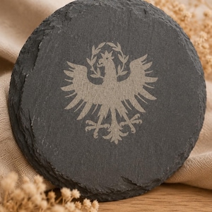 Könnte beinhalten: Ein runder Untersetzer aus dunkelgrauem Schiefer mit einem rauen, unebenen Rand. In der Mitte befindet sich eine hellgraue Gravur eines heraldischen Adlers mit ausgebreiteten Flügeln, unter einer kleinen Krone und einem Lorbeerkranz.