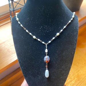 May include: A delicate necklace with a pearl chain and a pendant featuring a speckled blue, orange, and white bead, a small brown bead, and a teardrop-shaped pearl. The necklace is displayed on a black velvet bust.