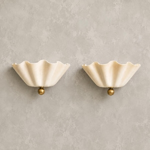 May include: A pair of wall sconces featuring off-white, scalloped, shell-shaped shades. Each fixture is mounted with a small, spherical, brass-toned metal accent at the base, providing a clean and symmetrical design for interior wall lighting.