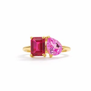 Puede incluir: Un anillo de oro con dos piedras preciosas prominentes colocadas una al lado de la otra. A la izquierda hay una piedra de color rojo intenso con corte esmeralda rectangular, y a la derecha una piedra de color rosa claro en forma de pera.