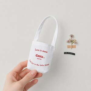 White miniature canvas tote bag held by hand against a wall with small flower decor.