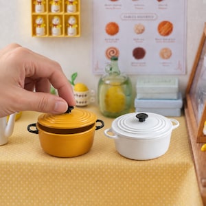 Puede incluir: Dos ollas miniatura redondas con tapa, una en color amarillo mostaza y otra en blanco. Cada olla cuenta con dos pequeñas asas laterales negras y un pomo negro en la tapa. Las ollas están colocadas sobre una superficie amarilla con lunares blancos.