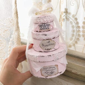 May include: A stack of three round, light pink boxes with distressed edges, decorated with white lace trim and fabric. The top box features a gold safety pin with pearls. Labels include the text 1871, Oh butterfly stay!, and Fabrique de Serrurerie.