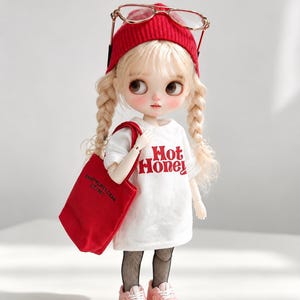 May include: A doll with blonde hair in two braids, wearing a red knit beanie, gold-rimmed glasses, a white t-shirt with the red text 'Hot Honey', a red tote bag, black fishnet stockings, and light pink sneakers with white laces.
