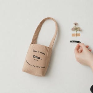 May include: A beige fabric bag with a single handle featuring the black printed text Less is more CHILL. SINCE 1950 and Find joy in the little things. The bag has a structured, paper-like texture and a simple, vertical rectangular shape.