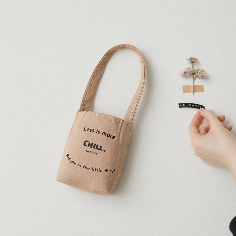 May include: A beige fabric bag with a single handle featuring the black printed text Less is more CHILL. SINCE 1950 and Find joy in the little things. The bag has a structured, paper-like texture and a simple, vertical rectangular shape.