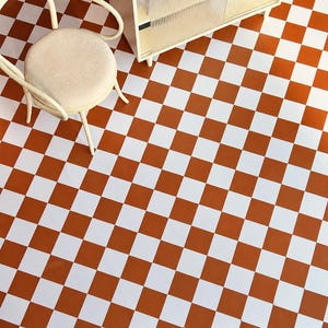 May include: A floor surface featuring a repeating pattern of alternating white and burnt orange square tiles. A light-colored wooden chair with a round, upholstered seat and a small cabinet with ribbed glass doors are placed on the floor.