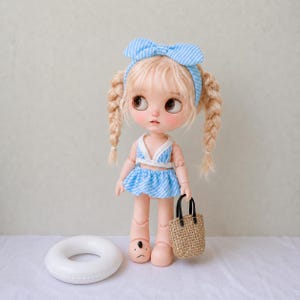 May include: A doll with blonde hair styled in two braids and a blue and white striped headband. The doll wears a blue and white striped bikini top and skirt, and light pink shoes with a black bear face design. It holds a small woven tan bag.